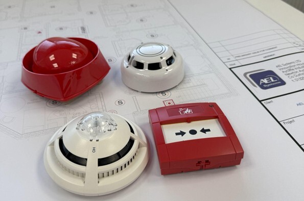 Selection of commercial fire alarm components including a smoke detector, sounder and manual call point laid out on building plans, representing system upgrades and modernisation for improved fire safety compliance.